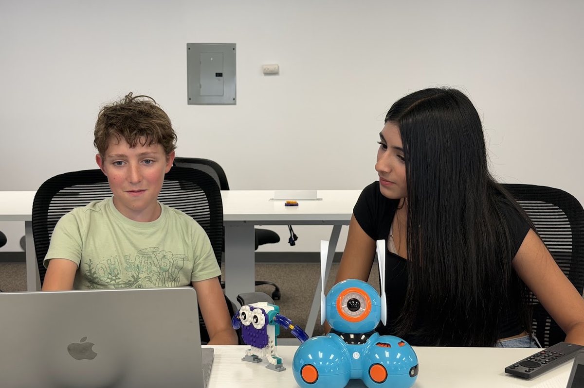 A student coding at a laptop with an instructor, next to a Dash robot and a LEGO robot